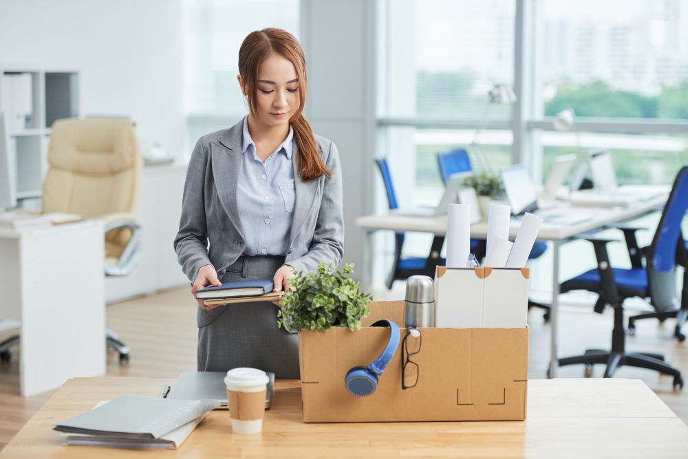 asian woman standing at desk in office with belongings in cardboard box